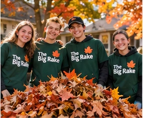 Four student volunteers smile together behind a freshly raked pile of golden leaves.